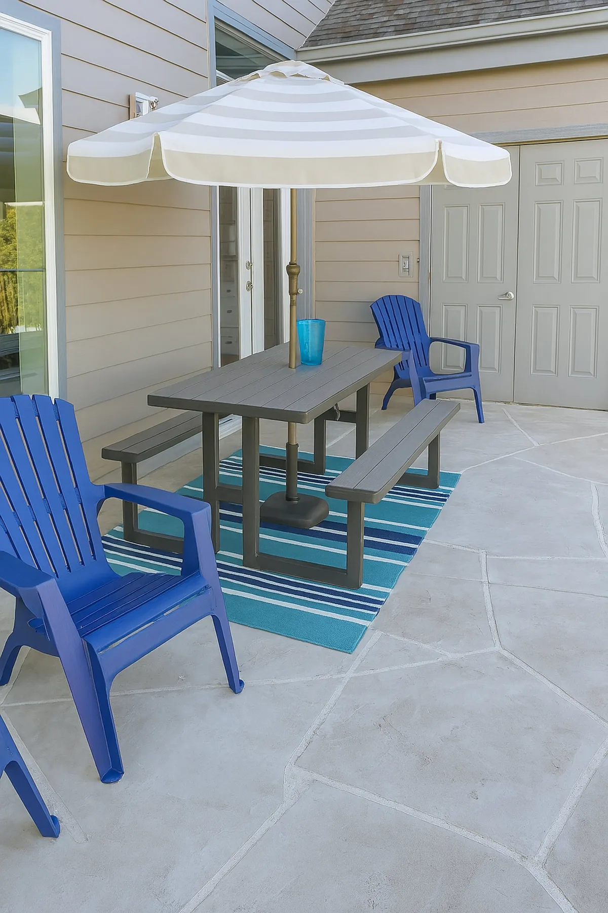 The image shows a cheerful outdoor dining area with blue Adirondack chairs, a striped umbrella providing shade, and a dining table set for a gathering. The setting exudes a lively and welcoming atmosphere, ideal for outdoor dining, relaxing, and enjoying special moments with loved ones.