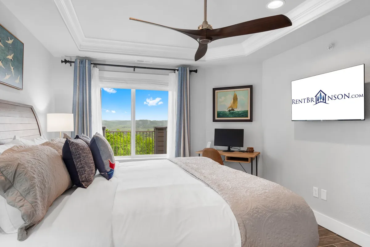 A stylish bedroom with a king bed adorned in decorative pillows, a spacious window offering a picturesque view, framed artwork, and a wall-mounted television for added comfort.