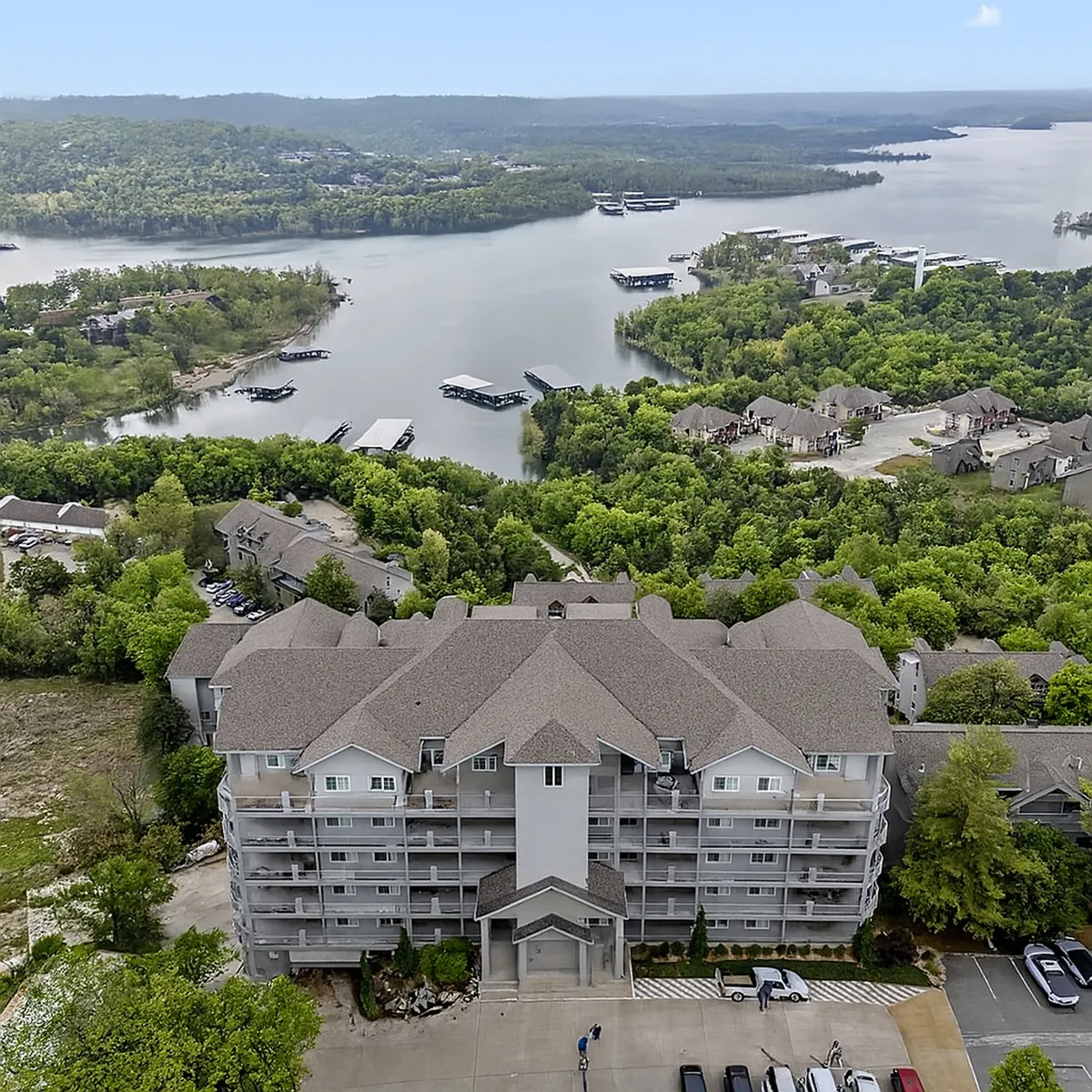 Taken from an exterior angle, this image highlights the multi-story building surrounded by lush greenery and showcases the proximity to a body of water with docks. The tranquil mood and the visible balconies suggest the presence of private outdoor spaces overlooking the lake.