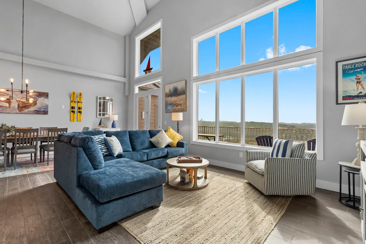 A comfortable living room setting with a plush sectional, dining area under a sparkling chandelier, expansive windows framing scenic views, decorative artwork, and a striped armchair. The mood is both inviting and luxurious, with plenty of natural light illuminating the space.